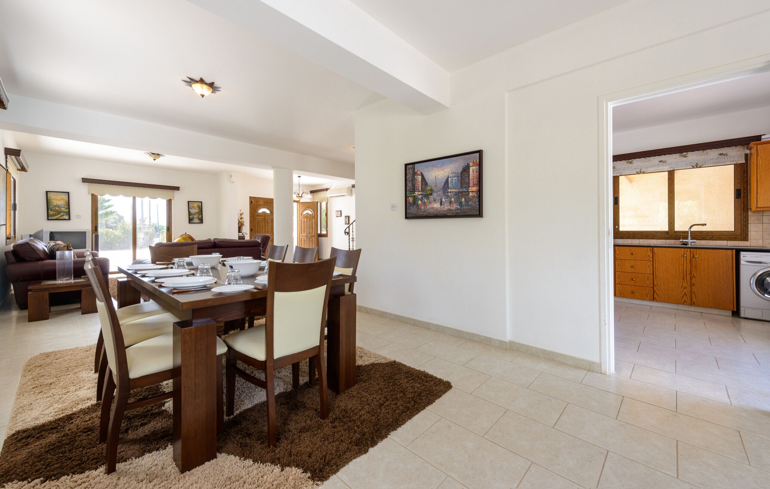 A spacious living area with a dining table, chairs, and a sofa. There is a kitchen area with wooden cabinets and a window.