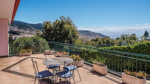 Ein Balkon mit Tisch und Stühlen, Blumentöpfen und Blick auf Stadt und Berge.