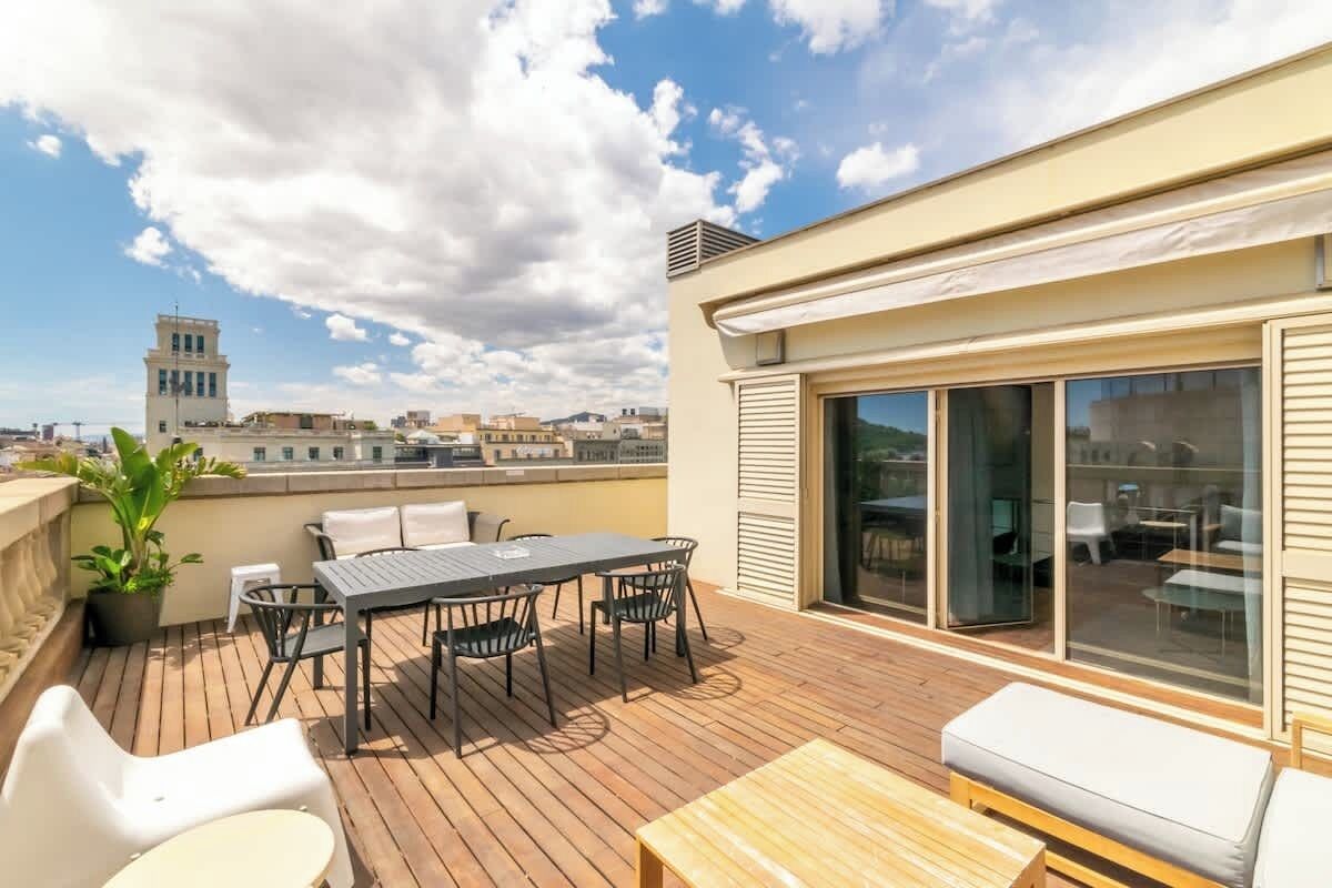 A rooftop terrace with a wooden deck, a table with chairs, a potted plant, and a view of the cityscape.