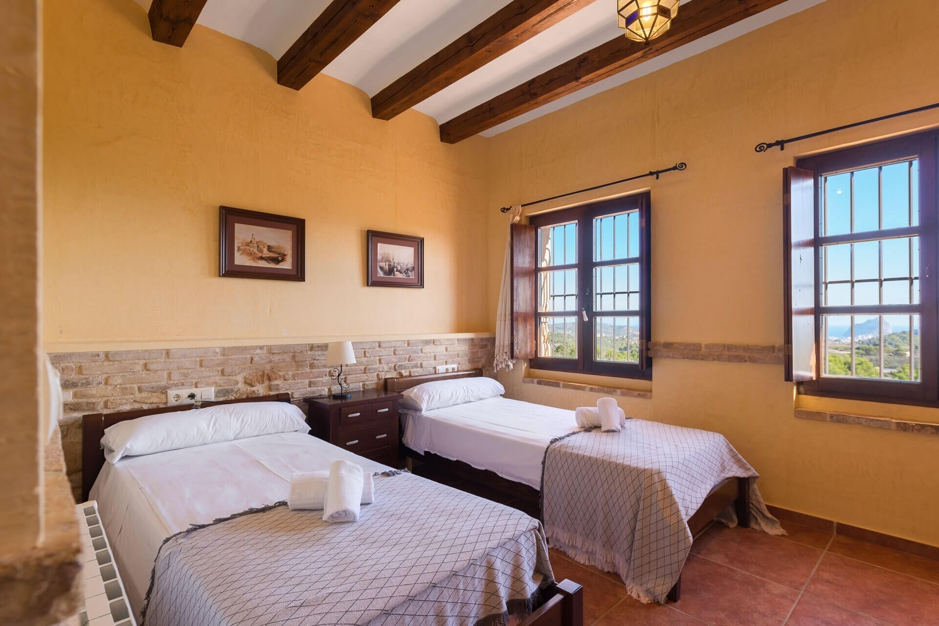 A room with two beds, a brick wall, wooden beams, and large windows with a view.