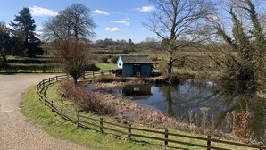A small blue building near a pond with a wooden fence and trees. - The Granary at Partridge Lodge (Bredfield, Woodbridge)