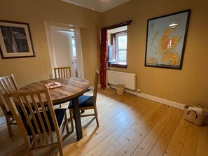 A dining area with a wooden table and chairs, a window with red curtains, a radiator, and a map of Scotland on the wall. - Cozy, dog friendly cottage on the edge of Kinblethmont Estate near to golf and beaches (By Arbroath)