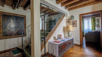 Una scala con ringhiera in vetro conduce a un piano superiore, con soffitto in legno, un quadro incorniciato, un tavolo con una lampada e un divano nella stanza adiacente.