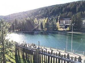 Vista de um lago com uma cerca de madeira ao primeiro plano, rodeado por árvores e edifícios ao fundo.