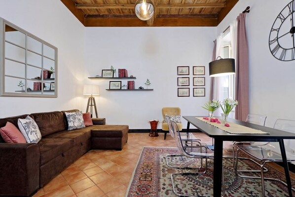 A living room with a brown sectional sofa, a dining table with chairs, a lamp, a clock, and a window with curtains.