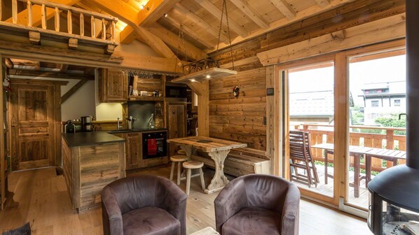 A rustic cabin interior with a wooden kitchen, dining area, and a balcony with outdoor seating.