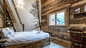 Una habitación de cabaña rústica con una cama, una silla de madera, un ventanal con vistas y un estante en la pared con libros.