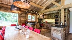 Um interior rústico em madeira com uma grande mesa de jantar, cadeiras estofadas em vermelho, uma área de cozinha com armários de madeira e um lustre pendurado.