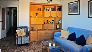 A living room with a blue sofa, a wooden bookshelf, a patterned armchair, a glass coffee table, and a ceiling fan. - Sunrise watching Tabarca (Santa Pola)