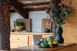 A rustic kitchen with wooden beams, a wooden cutting board, a black teapot, and a vase with flowers on the counter. - Altana studio (Venezia)