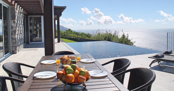 Un tavolo da pranzo con una ciotola di frutta, piatti e posate, con vista su una piscina e l'oceano.