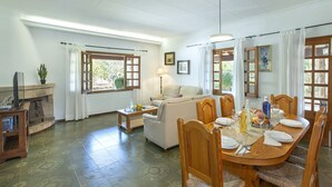 Uma sala de jantar com uma mesa de madeira posta para quatro pessoas, um sofá e uma televisão no canto.