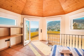 Quarto com teto de madeira, cama com manta listrada, vista para uma cidade costeira e varanda com grade.
