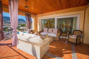 A balcony with wicker furniture, a glass door leading to a pool area, and a view of a mountainous landscape.
