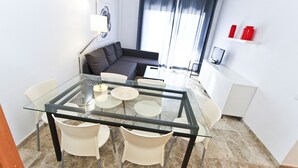 Un salon moderne avec une table à manger en verre, des chaises blanches, un canapé gris et un téléviseur posé sur une étagère blanche.