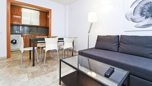 Un salon moderne avec un canapé gris, une table basse en verre et une télécommande posée dessus. On y trouve également un coin repas avec une table et des chaises, ainsi qu'une cuisine dotée de meubles en bois et d'un lave-linge.