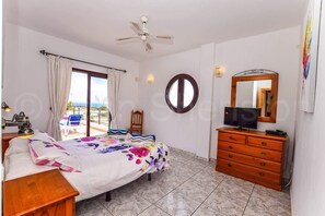 A bedroom with a bed, a dresser, a ceiling fan, a TV, a mirror, and a view of the outdoors.