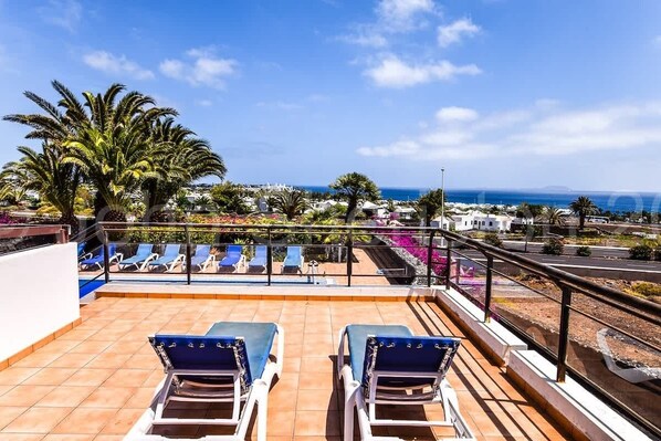 A rooftop terrace with sun loungers, palm trees, and a view of the sea and buildings. - Villa Montana - Large family Villa by Villas Now (Playa Blanca)
