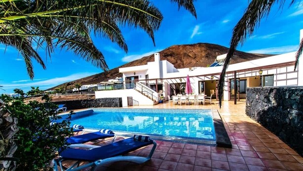 A pool area with lounge chairs, a stone wall, and a building with a balcony.