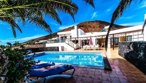 A pool area with lounge chairs, a stone wall, and a building with a balcony. - Villa Montana - Large family Villa by Villas Now (Playa Blanca)