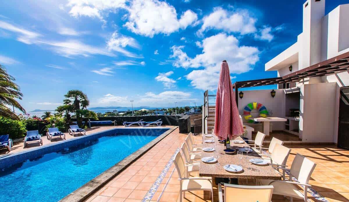 A poolside area with a dining table set for two, lounge chairs, and a covered umbrella.