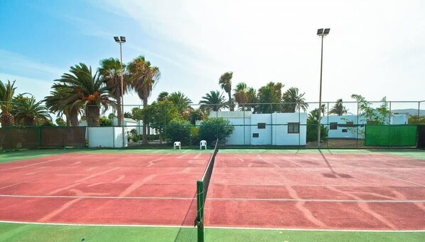 An outdoor tennis court with a net, surrounded by a fence, palm trees, and buildings in the background. - Las Brisas 112 - Resort Stay by Villas Now (Playa Blanca)