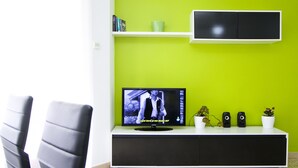 Een moderne woonkamer met een flatscreen-tv, een witte tv-meubel, twee zwarte stoelen, een groene muur en een potplant.