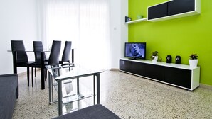 Een moderne woonkamer met een glazen eettafel, zwarte stoelen, een flatscreen-televisie, een witte televisiemeubel en een groene accentmuur.