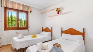 A hotel room with two single beds, a window with a view of trees, a small shelf with a vase, and a door with a patterned curtain. - Villa Cruz - PlusHolidays (Calpe)