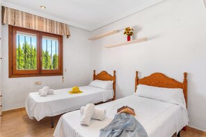 A hotel room with two single beds, a window with a view of trees, a small shelf with a vase, and a door with a patterned curtain. - Villa Cruz - PlusHolidays (Calpe)