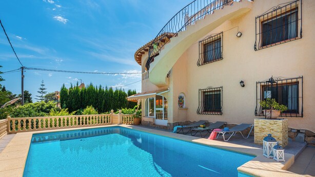 A swimming pool with a tiled edge, a covered patio area with lounge chairs, and a building with multiple windows and balconies. - Villa Cruz - PlusHolidays (Calpe)