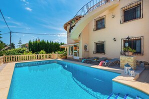 A swimming pool with a tiled edge, a covered patio area with lounge chairs, and a building with multiple windows and balconies. - Villa Cruz - PlusHolidays (Calpe)