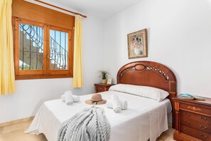 A bedroom with a wooden bed, a bedside table, a nightstand, a window with curtains, a framed picture on the wall, and a potted plant. - Villa Cruz - PlusHolidays (Calpe)