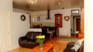 A living room with a brown leather sofa, a wooden coffee table with a vase of yellow flowers, a chandelier, and a kitchen in the background.