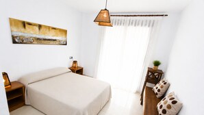 Een slaapkamer met een bed, nachtkastjes, een hanglamp, een raam met gordijnen, een houten bankje en een bloempot met plant.