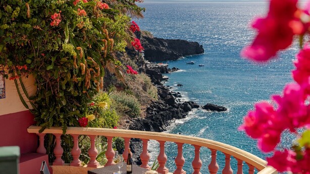 A balcony with a table, chairs, and a bottle of wine, overlooking a rocky coastline and the sea.