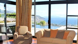 A room with a sofa and armchair facing a large window with a view of a coastal landscape. - Fabulous seaview, pool and games room | Villa Luz (Funchal)