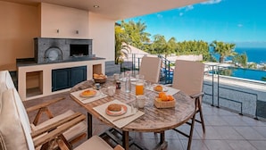 A dining area with a table set for breakfast, a stone fireplace, and a view of the ocean. - Fabulous seaview, pool and games room | Villa Luz (Funchal)