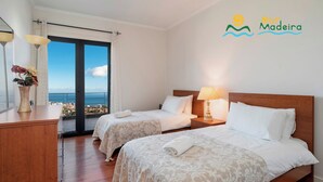 A hotel room with two beds, a wooden dresser, a vase with flowers, and a view of the sea. - Fabulous seaview, pool and games room | Villa Luz (Funchal)