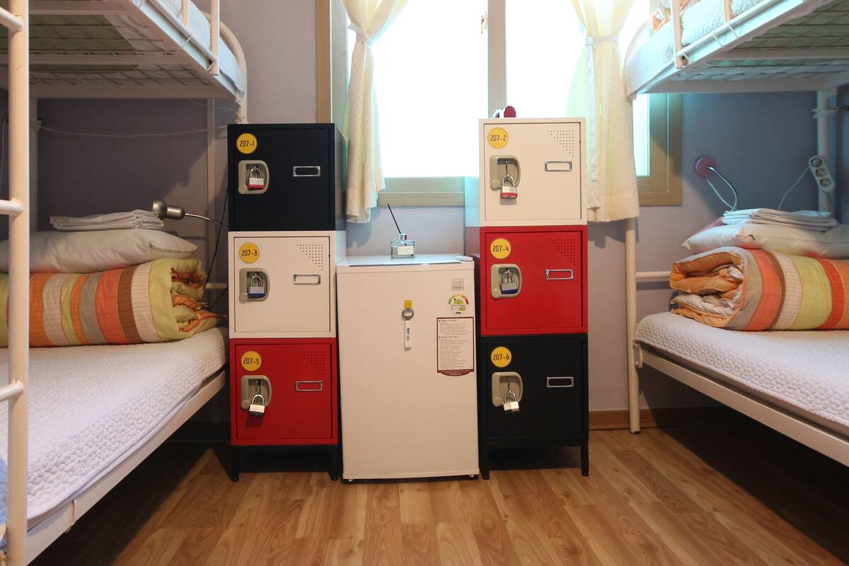 A dormitory room with bunk beds, a desk with a computer, and a small refrigerator.