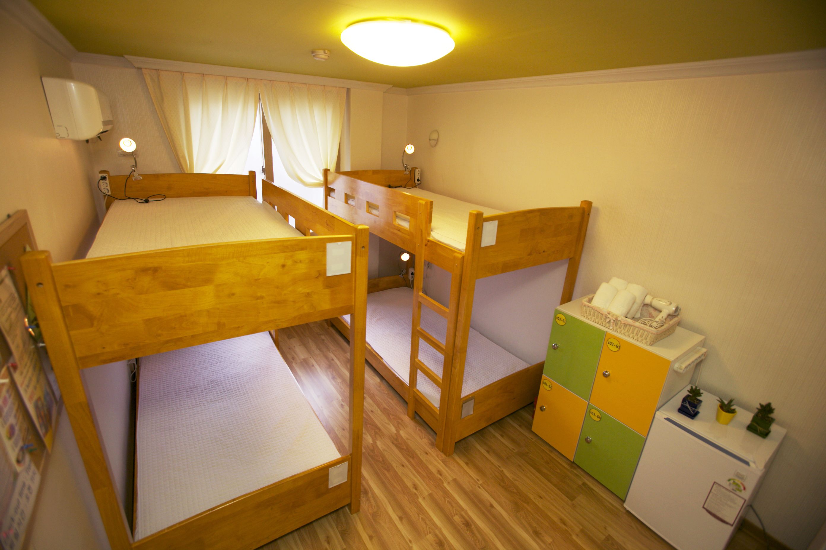 Een kamer met stapelbedden, een klein kastje met een koelkast, en een raam met gordijnen.