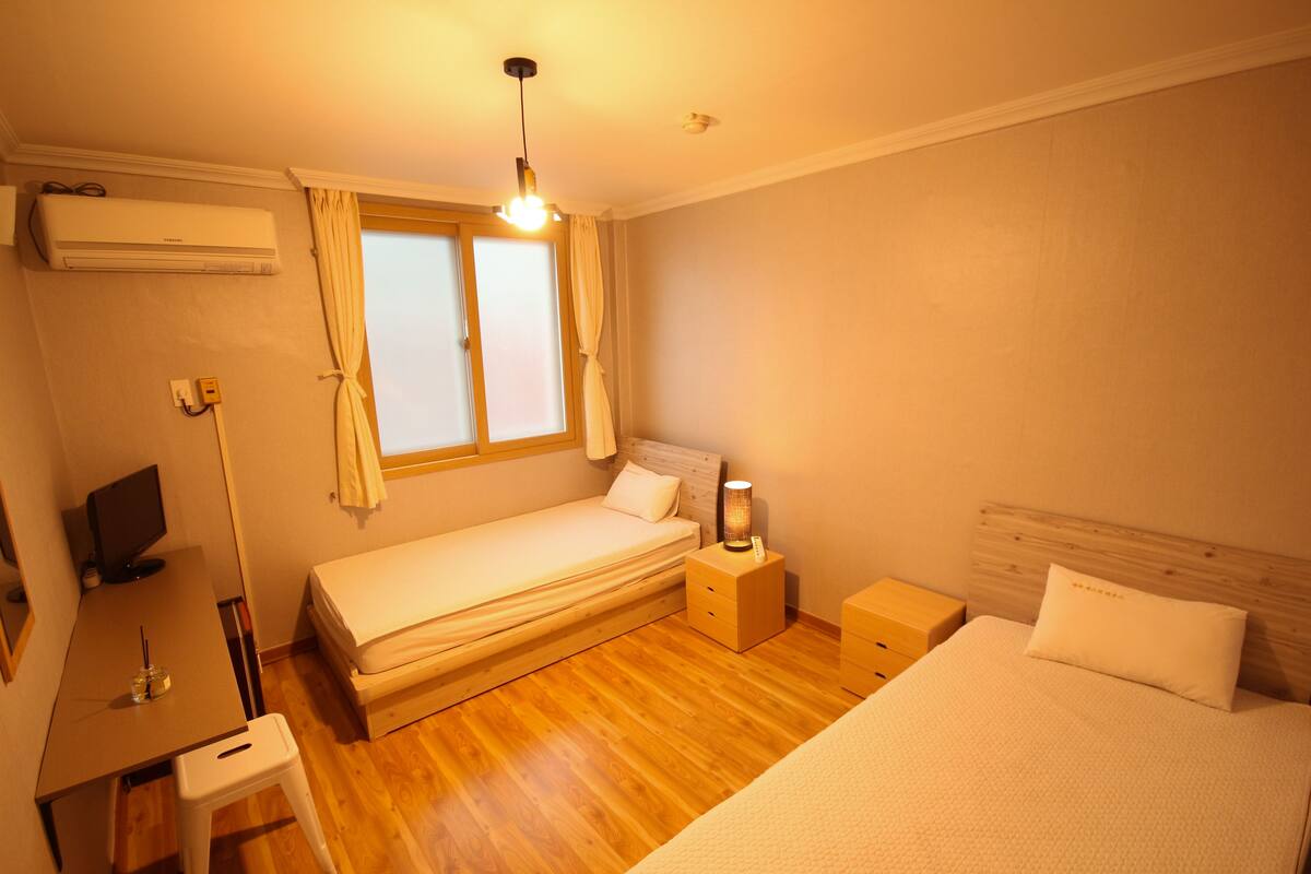 A compact bedroom with a single bed, a desk with a computer, a bedside table with a lamp, and an air conditioning unit on the wall.