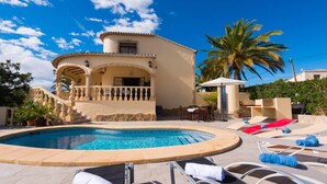 Un espace aménagé au bord de la piscine, avec des chaises longues, un palmier et un bâtiment doté d’un balcon.