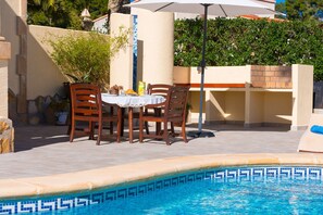 Un área de piscina al aire libre con borde de azulejos, una mesa con sillas y una planta en maceta.