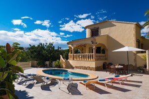 A pool area with lounge chairs, a building with a balcony, and a clear blue sky.