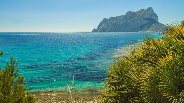Un paysage côtier caractérisé par des eaux turquoise limpides, une montagne rocheuse imposante et une végétation luxuriante au premier plan.