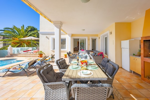 A covered patio with a dining table set for six, surrounded by wicker chairs, a pool, and a kitchen area with a refrigerator.