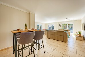 A spacious living area with a dining table, two bar stools, a sofa, and a television mounted on the wall.