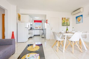 A modern kitchen with white cabinets, a dining area with a white table and chairs, and a living room with a gray sofa.