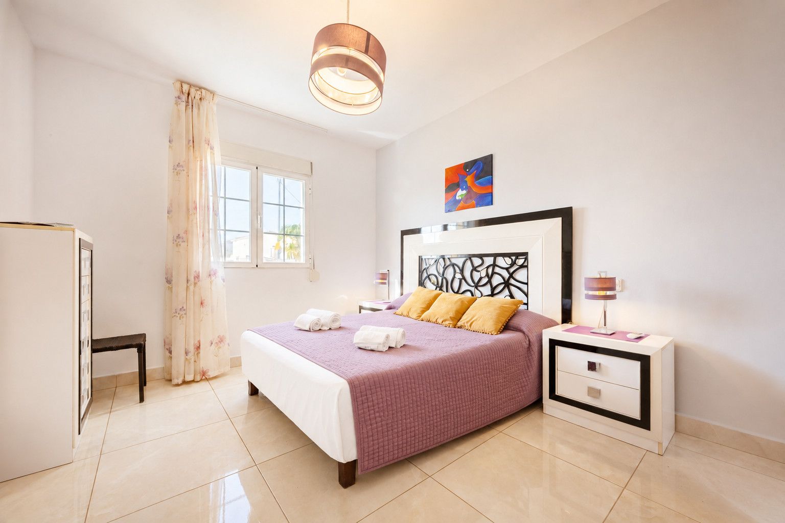 Habitación de hotel con cama, mesitas de noche, escritorio, silla, ventana con cortinas y un cuadro en la pared.